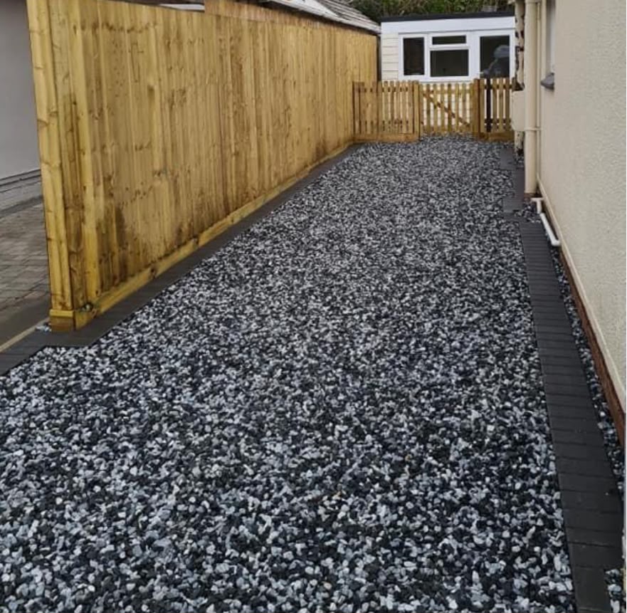 Black basalt decorative gravel side driveway with block paving edging beside a fenced boundary